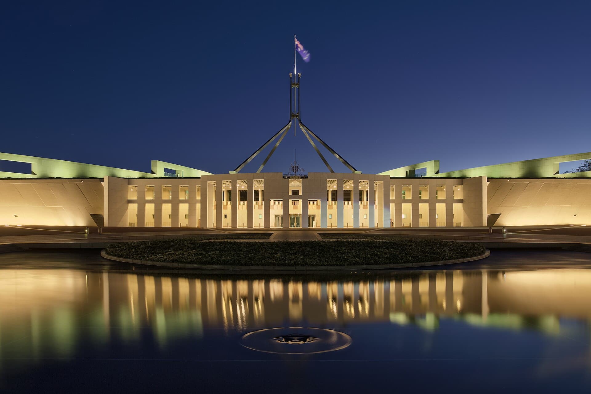 Parliament House in Canberra at dusk, lights on beneath the flag.